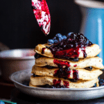 stack of pancakes topped with blueberries and drizzled with fruit sauce