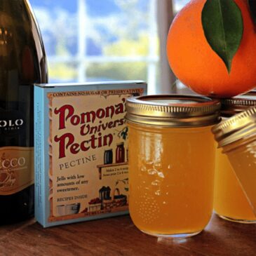a bottle of champagne, next to a box of Pomona's Pectin, jelly jars and a fresh orange