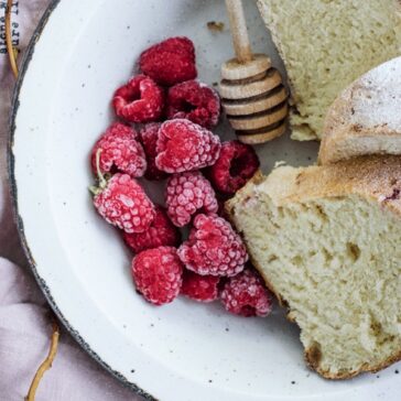 fresh raspberries on a white plate next to fresh bread and a honey comb