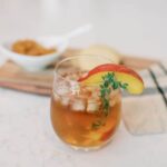 glass on a counter top with a slice of peach and sprig of. thyme next to a bowl of State-Line Jam